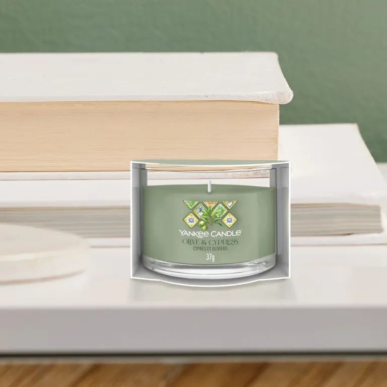 Candela verde in vetro e confezione. Scritta: Yankee Candle, Olive & Cyprès, 37g. Su tavolo con libri.