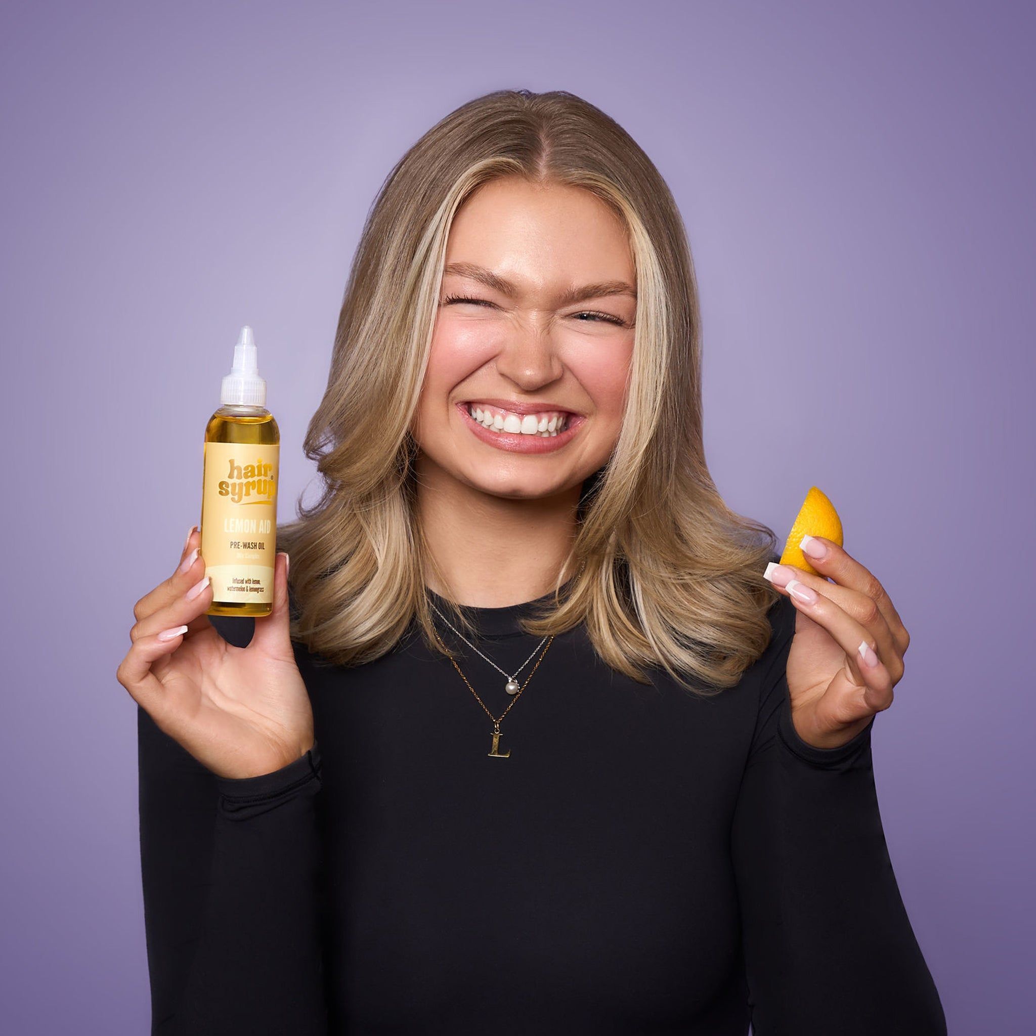 Donna con flacone "Hair Syrup Vitamin C Me" e fetta d'arancia. Pre-Wash Oil. Sfondo viola.