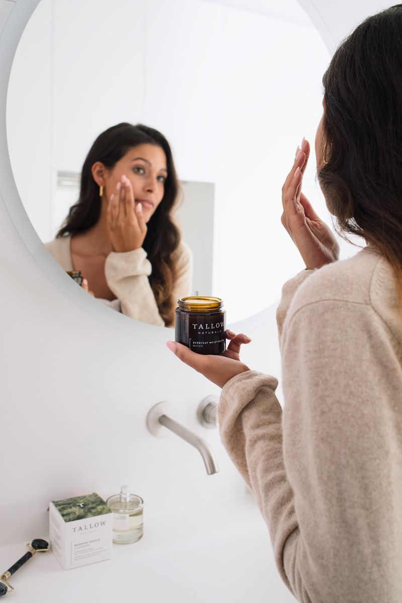 Donna si guarda allo specchio. Tiene un vasetto. Scritta: Tallow Naturals, Everyday Moisturizer Natur. Altri prodotti visibili.