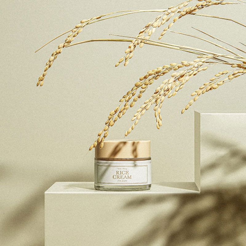 Vasetto di vetro con coperchio beige. Scritta: Rice Cream, I'M FROM. Decorato con spighe di riso. Su piedistallo bianco.