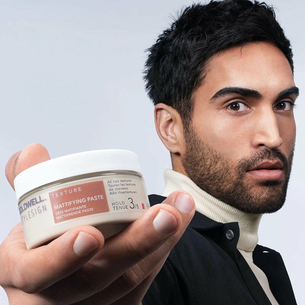 Uomo tiene Goldwell Stylesign Texture Mattifying Paste. Nome e marchio visibili. Coperchio bianco, barattolo beige.