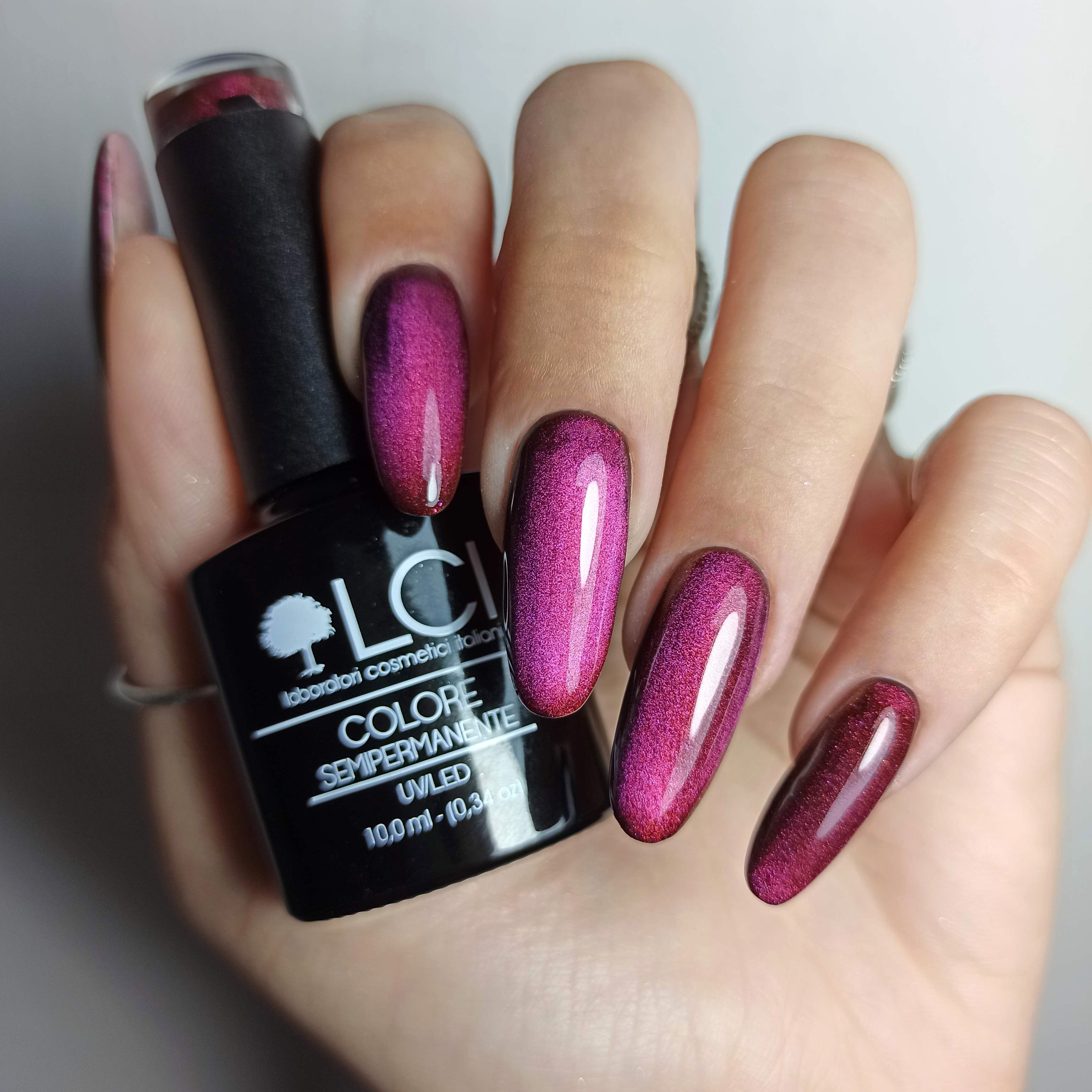 Mano con unghie smaltate e flacone di smalto. Unghie rosa-rosso. Scritta: LCI, COLORE SEMIPERMANENTE, UVLED 10,0 ml.