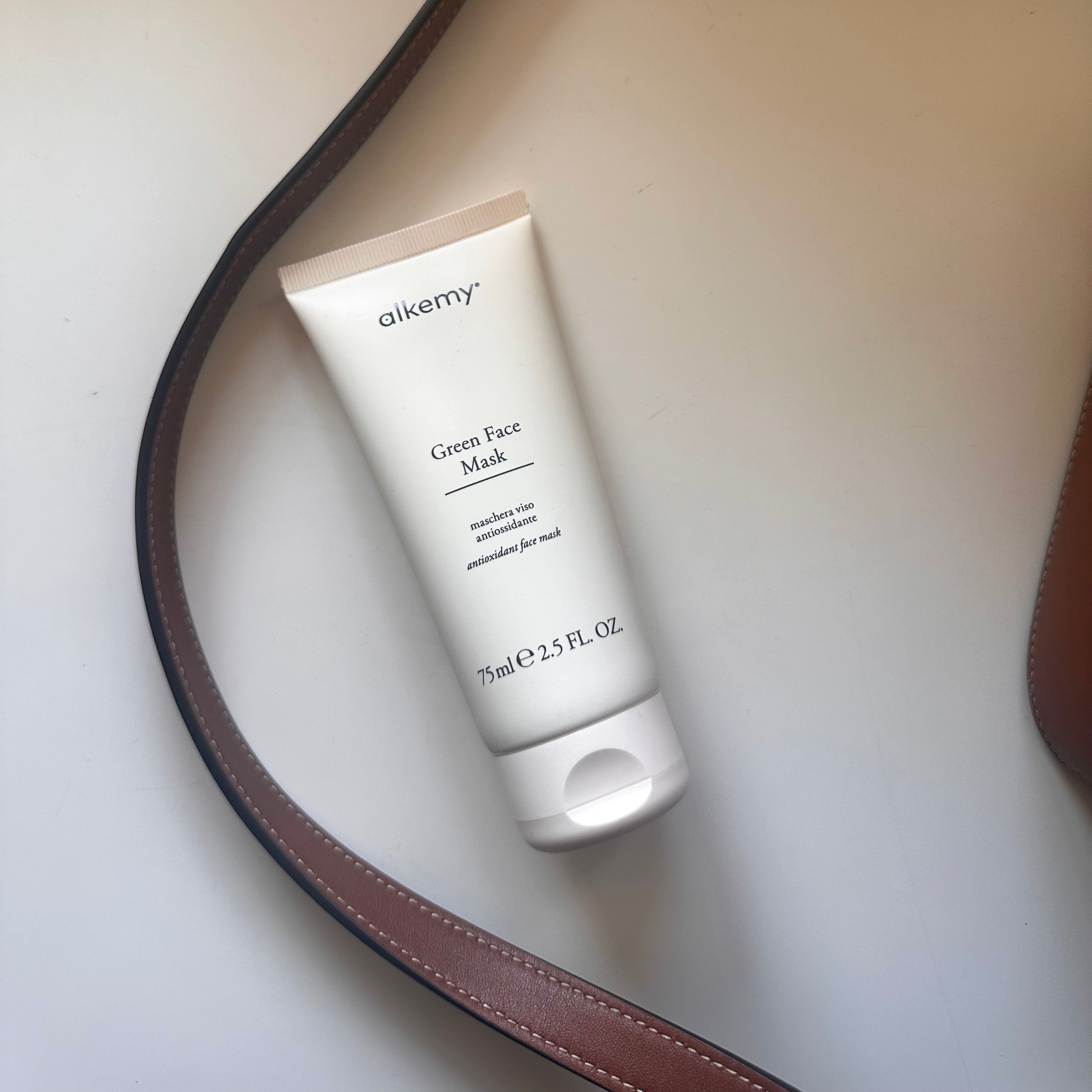 Tubo bianco Alkemy Green Face Mask accanto a una borsa in pelle marrone.
