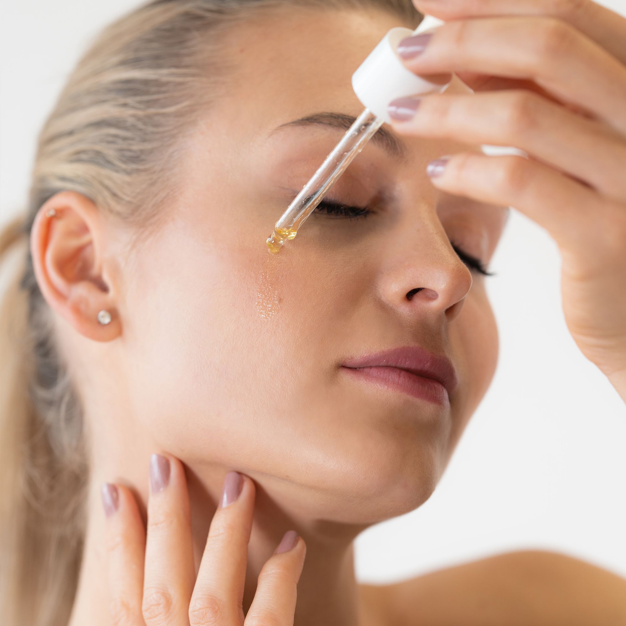 Donna con occhi chiusi applica olio sul viso con contagocce. Pelle chiara, capelli chiari.