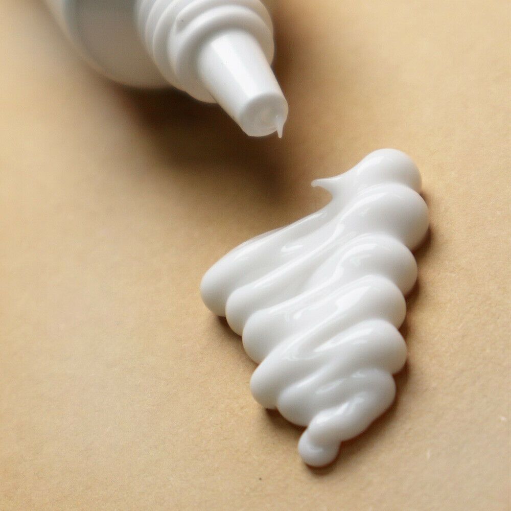 Primo piano di crema bianca che esce da un tubetto. La crema è distribuita su una superficie beige chiaro.