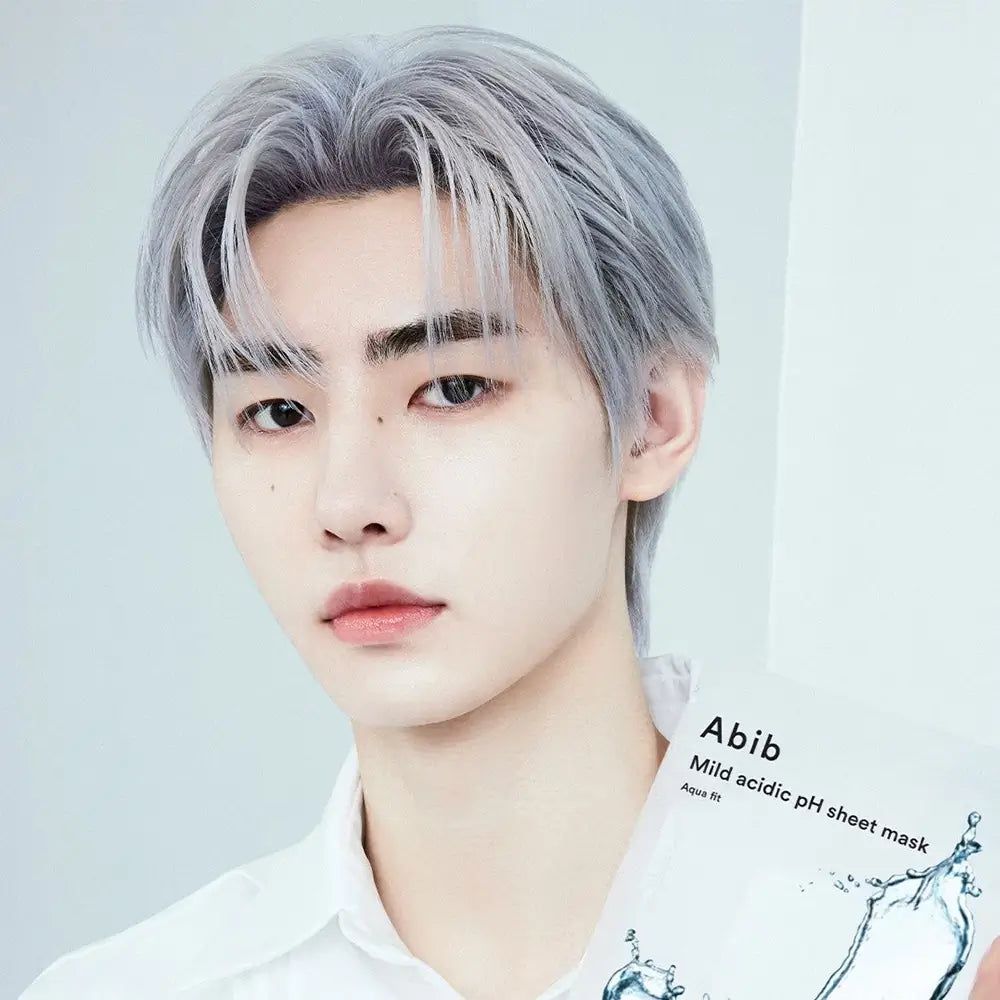 Persona che tiene la maschera viso ABIB Milde Säure pH Aqua Fit. Confezione del prodotto con il nome del prodotto. Persona con capelli grigi e camicia chiara.